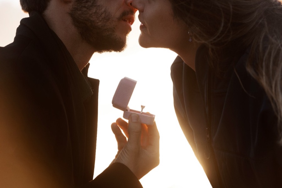Pedida de mano al atardecer: pareja muy cerca y anillo de compromiso en un estuche rosa.